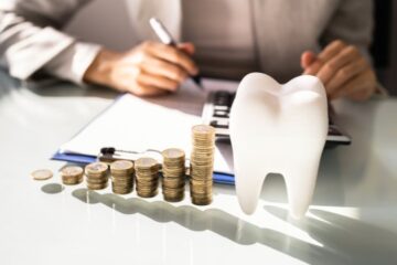 Stapel von Münzen vor einem Zahnmodell, im Hintergrund eine Person mit Taschenrechner – Symbolbild für die Finanzierung und Kostenplanung in einer Zahnarztpraxis.