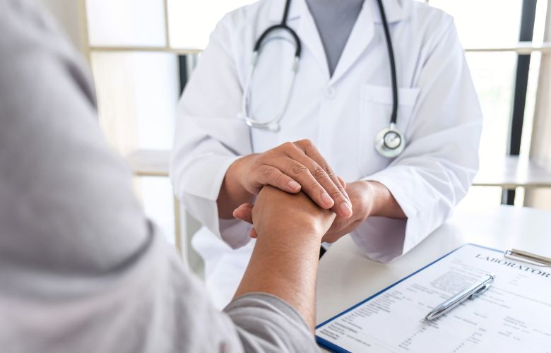 Arzt hält die Hand eines Patienten in empathischem Gespräch mit Laborbefund auf dem Tisch